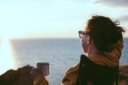 Back view of young woman traveler holding iron mug cup with tea or coffee, enjoying sunset scenery view of sea landscape, sitting in touristic chair. Travel camping and adventure lifestyle concept