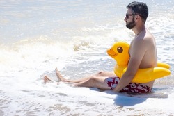 Upset brunette young man in sunglasses sitting on sandy beach near blue sea, wearing yellow duck inflatable ring. Sad facial expression. Feeling fear, phobia of water, I can not swim. Copy space.