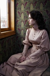 Young woman in beige vintage dress of early 20th century looking trough the window in coupe of retro railway train
