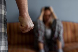 Photo of young woman sitting on sofa at home,focus is on man's fist in the foreground of the image.Home violence concept.Frightened woman and men's fist.Woman is victim of domestic violence and abuse.