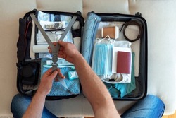 Happy traveler packing his clothes into a suitcase, getting ready for a trip. Man packing passport, face masks and vaccination certificate. Themes personal protection and flight rules during COVID-19.