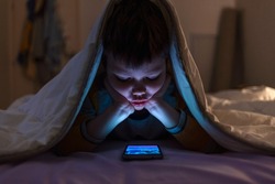Little boy on cell phone under blanket. Curious boy sits in bed under white blanket and play games on smartphone in the dark room.  Image of nice boy in pajama sitting in front of mobile. 