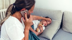 Worried mother checking temperature of her little son who is lying in bed with fever. Young mother checking the temperature of her ill 6 years old boy on a couch in the living room, calling doctor.