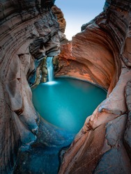Beautiful turquoise waterfall flows into natural pool surrounded by red rock formation in Karijini National Park in Western Australia.