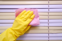 Women's hands in yellow gloves wash off the dirt from the yellow-pink blinds on the window with a pink rag, close-up