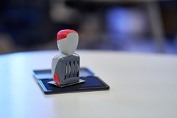Office Stamp with Four Rolling Wheels, Resting on an Open Stamp Pad Filled with Blue Ink. Red Details on the Stamp Head and Body. White, Curved Office Desk. Blurred Background, Window Reflections