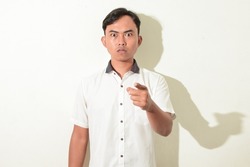portrait of asian man glaring and pointing at the camera. Indonesian man poses angry, glaring, and intimidating. asian man in white shirt isolated on white background

