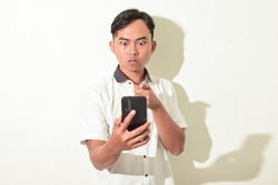 portrait of asian man communicating video call using smartphone. Indonesian man poses angry, glaring, and intimidating while staring at the cellphone screen. asian man in white shirt isolated on white