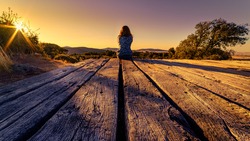 105 / 5000
Woman from behind sitting on a wooden floor, contemplating the horizon with sunset in the field.