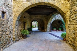Picturesque alley with stone houses and arched passageway with green plants on the ground, Monells, Girona, Spain