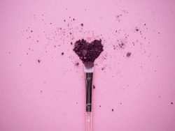 Crushed plum colored shimmer eyeshadows for smokey ice effect scattered in heart form on pink background with nylon makeup brush top view. 
