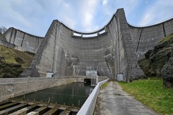 An impressive dam on a river in France showing the water supply channel and the dam wall during the dry season with blue sky above and green grass and rocks to either side
