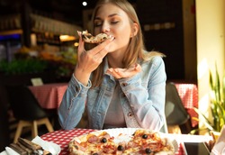 Funny blonde girl in jeans jacket eating pizza at restaurant.