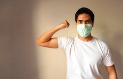 Portrait of a handsome Asian man wearing medical protective mask standing with a raised arm shows strong biceps. Isolated background. Concept of staying strong and healthy against coronavirus covid-19