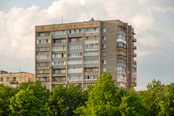 Typical facade of an old brick nine-story building in the post-Soviet countries on a background of green trees.