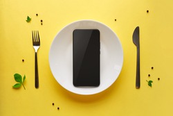 Smartphone on white plate with fork and knife. Food delivery concept on yellow background