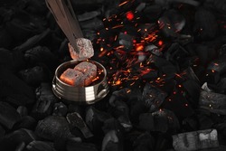 With hookah tongs, they shift and blow on burning cubes of coconut charcoal in a metal kaloud, sparks fly on a black background. Horizontal orientation, close-up, focus in the foreground, no people.