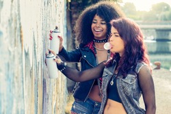 two urban teenage girls painting graffiti artwork on a city wall. young trendy afro-hair girl with girlfriend drawing on wall with spray can and chewing bubble gum. modern youth culture concept.