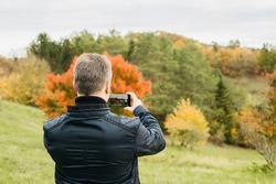 A man takes a photo of the autumn landscape on a mobile phone. Mobile photography as a hobby. Activity in nature. Blogging