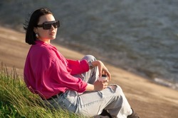 Brunette woman in sunglasses sitting on lakeshore and chilling in the sun rays outdoor