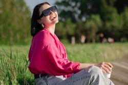 Brunette woman in sunglasses sitting on lakeshore and chilling in the sun rays outdoor