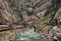 Train riding deep in the royal gorge beside the arkansas river in Colorado. Orange engine beside rushing blue water with rocky cliffs all around