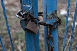 An old rusty metal castle and a large iron chain at a dilapidated gate with shabby blue paint on a pale background in muted horror-style tones, a locked wicket leading to an abandoned site.