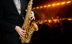 Male Musician in a Formal Black Suit Holds a Tenor Saxophone on the background of the concert atmosphere. Saxophonist Plays Jazz.  Saxophone Close-up. Copy space.                           