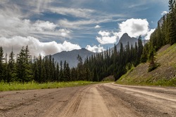 A dry dirt road under, a cloudy blue sky, leading to a forest below some rocky mountains.