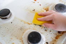 A hand with a yellow wash sponge washes the very dirty greasy surface of the gas stove. After the sponge, a clean trace remains