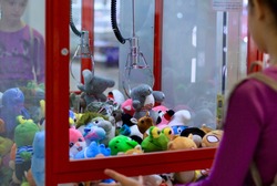 Caucasian girl playing toy crane vending machine, Claw Game or Cabinet to Catch the Toys. Shopping, holiday activity, game of chance, vacation concept. Selective focus on crane.