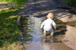 Little two year old boy running around the puddles. Cute child wearing a grey jumpsuit and brown leather boots. Concept of happy childhood. 