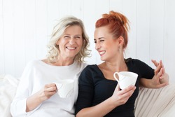 Two beautiful women-a mother and daughter sitting on the sofa at home, snuggled. They drink coffee, tea. Relationships and shared time.