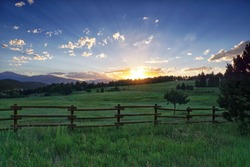 Sun rays streak the sky over a lush green meadow as the setting sun slips beyond the horizon. This image was captured on a warm summer evening in the foothills of the Rocky Mountains near Genesee, CO.