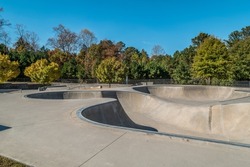 Empty skateboard course made of cement with metal railing and lots of twists and turns going over humps into the deep pool with the colorful woodlands in the background in autumn