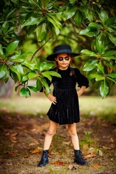 a small, beautiful girl in a black dress and hat, a face in Halloween makeup, stands near a tree in the park