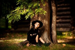 a little girl, in a black dress and hat,sits under a tree in the park and holds a bat, a toy mouse, in her hands, on Halloween