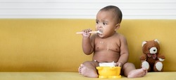 Adorable African baby newborn in diaper sitting on sofa with small bear doll trying to grab a spoon to feed herself. Spilled food messy in mouth and body of little kid. Child development concept