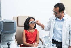Happy Indian child girl doing examine eyesight with trial frame and lens with ophthalmologist or optometrist for eyes test at hospital or optometry clinic