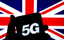 5G letters on a silhouette of a smartphone in the hand with the blurred United Kingdom flag on the background. Authentic photo, not a montage or illustration. 