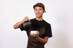 Handsome barista asian man wearing brown apron and black t-shirt isolated over white background . Barista holding milk jug and coffe cup practicing making coffee latte