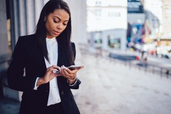 African American female entrepreneur in corporate suit using online banking for transferring money distantly via smartphone. Confident businesswoman texting email letter on cellphone using internet