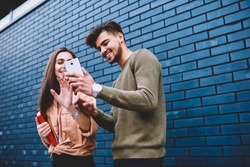 Cheerful best friends viewing funny photos in social networks via smartphone standing together on street, happy male and female hipsters watching funny online video content on mobile phone outdoors