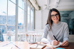 Cheerful hipster girl working as journalist enjoying leisure at cafeteria and looking at camera during break, successful happy writer creating new novel ideas while sitting at coworking space
