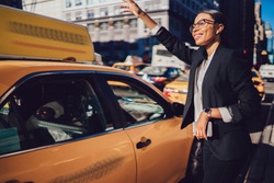 Cheerful afro american manager in formal wear catching yellow taxi for getting to work standing in traffic movement, happy female lawyer in elegant clothes waving hand on street of modern metropolis