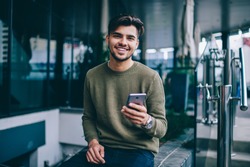 Portrait of cheerful handsome male using mobile phone and wireless internet for chatting and networking , young hipster guy looking at camera satisfied with good 4G connection for sending messages