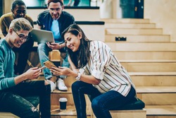 Cheerful millenial woman showing latest feed from social networks to group of multiracial friends, happy crew of male and female colleagues spending free time in college talking and using gadgets