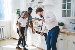 Professional bearded chubby man in formal outfit with clipboard inspecting oven together with attentive ethnic couple while standing at modern sunny apartment