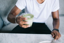 Faceless ethnic hipster black man in casual white T shirt holding glass of healthy fresh green smoothie sitting on sofa 