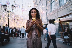 Attractive female tourist in optical glasses for stylish look walking in evening city during solo travelling, Asian millennial user in bluetooth headphones watching online video via smartphone gadget